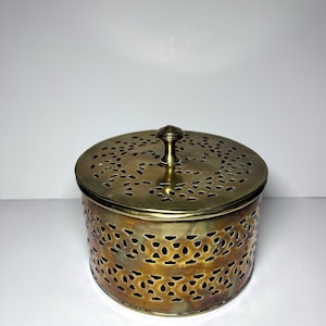 May include: A round, antique brass container with a lid. The lid and sides feature intricate cut-out patterns. A small, rounded handle sits atop the lid. The container is likely used for storage or decoration.