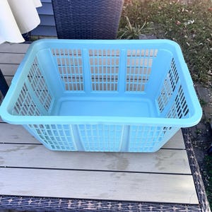 May include: A light blue rectangular laundry basket with a grid pattern. The basket is made of plastic and sits on a wooden deck. The basket has a rectangular shape with a slightly curved rim.