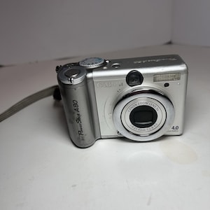 May include: Silver Canon PowerShot A80 digital camera with a Canon Zoom Lens 3x. The camera has a flash, a lens, and a strap. The camera is on a white surface.