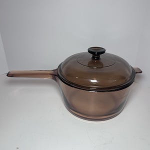 May include: A brown glass saucepan with a long handle and a matching lid. The saucepan has a rounded shape and is designed for stovetop cooking. The lid has a black knob. The cookware is made of transparent glass.