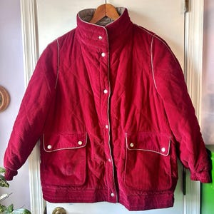 May include: A red corduroy jacket with a quilted design. The jacket has a high collar, white trim, and four front pockets. It features white buttons and a gray lining. The jacket is hanging on a wooden hanger.