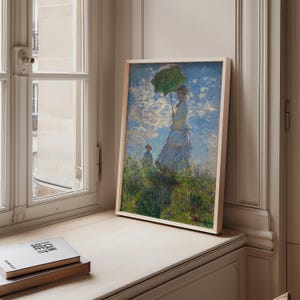 May include: A framed print of a classic Impressionist painting. The artwork depicts a woman with a green parasol standing in a field, with another figure in the background. The painting has a light wooden frame and is displayed on a windowsill.