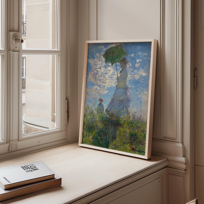 May include: A framed print of a classic Impressionist painting. The artwork depicts a woman with a green parasol standing in a field, with another figure in the background. The painting has a light wooden frame and is displayed on a windowsill.