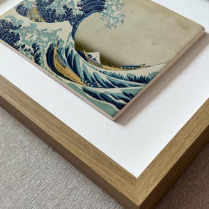 May include: A framed print featuring the iconic Great Wave off Kanagawa artwork, showcasing a large, curling blue wave with white foam crests and yellow boats, set against a muted beige background, enclosed in a light brown wooden frame.