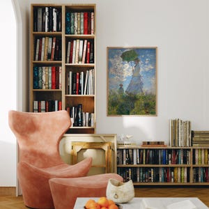 May include: A room interior featuring a framed artwork of a woman with a green umbrella, a pink armchair, and two wooden bookshelves filled with books. A matching ottoman sits in front of the chair. A marble coffee table holds a decorative bowl and oranges.