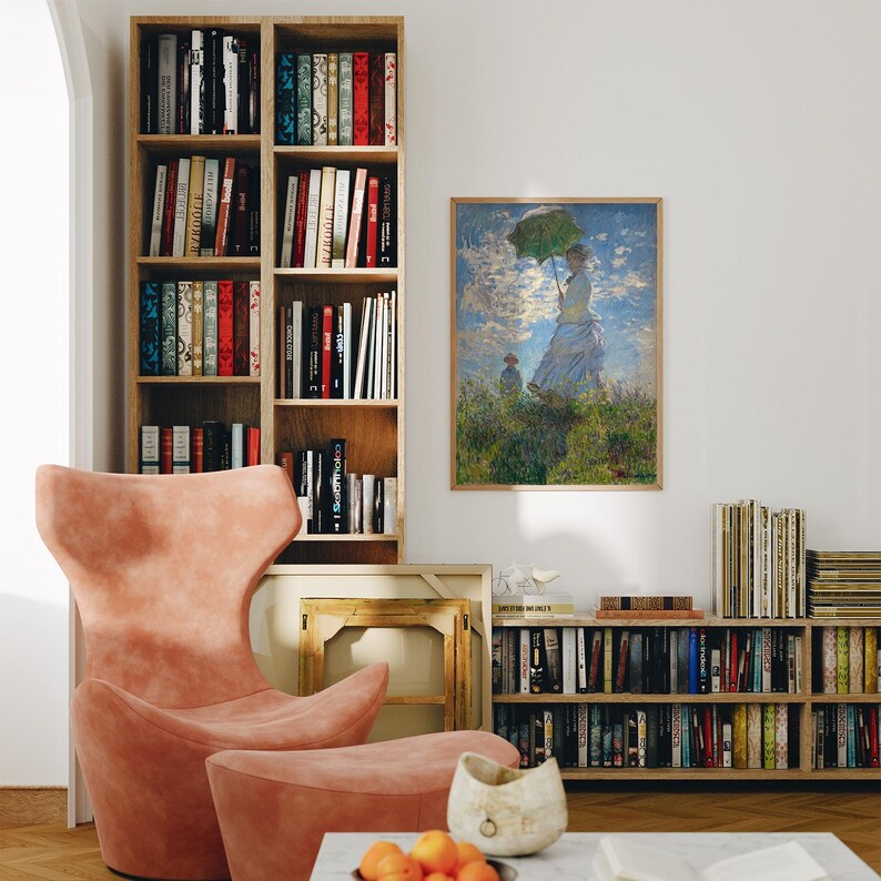 May include: A room interior featuring a framed artwork of a woman with a green umbrella, a pink armchair, and two wooden bookshelves filled with books. A matching ottoman sits in front of the chair. A marble coffee table holds a decorative bowl and oranges.