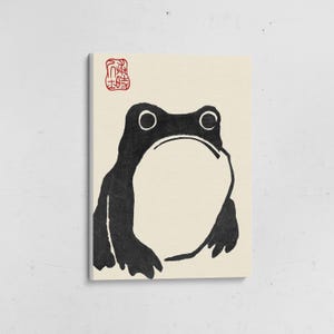 May include: A canvas print featuring a minimalist black and white illustration of a frog. The frog is depicted with a large open mouth and simple lines. A red stamp is in the upper left corner. The background is a cream color.