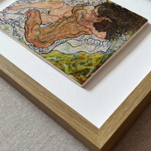 Peut inclure: Une impression d'art encadrée représentant une silhouette allongée aux cheveux sombres, réalisée avec des coups de pinceau expressifs dans des tons orange, marron et vert. L'œuvre est sur un passe-partout blanc dans un cadre en bois clair texturé.