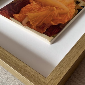 May include: A framed art print featuring a painting of a figure draped in a vibrant orange fabric, resting on deep red textiles. The artwork is set within a textured, metallic gold frame with a wide white mat border, showcasing a classic composition.