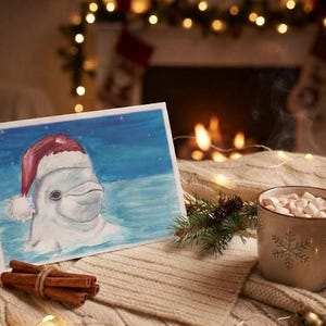 Pode incluir: Um cartão de felicitações com a pintura de uma baleia beluga branca usando um gorro de Pai Natal vermelho e branco sobre um fundo azul, ao lado de uma caneca bege com chocolate quente e marshmallows, um ramo de pinheiro e canela.
