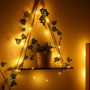 Może przedstawiać: Wisząca drewniana półka z małą rośliną doniczkową i dekoracyjnym bluszczem. Półka jest zawieszona na sznurze i ozdobiona lampkami choinkowymi, tworząc ciepłą atmosferę. Półka ma około 30 cm szerokości.