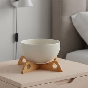 May include: A white ceramic bowl sits on a light brown bamboo stand, placed on a light wood nightstand. A white wall lamp is mounted on the wall behind the nightstand, and a bed with a beige headboard is in the background.
