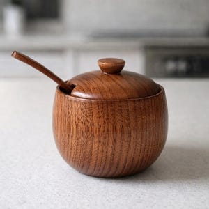 Könnte beinhalten: Eine kleine, runde Zuckerdose aus Holz mit Deckel und kleinem Löffel. Die Schüssel und der Löffel sind aus dunkelbraunem Holz mit sichtbarer Maserung gefertigt. Der Deckel hat einen kleinen Knopf oben. Der Löffel liegt in einem Schlitz an der Seite der Schüssel.