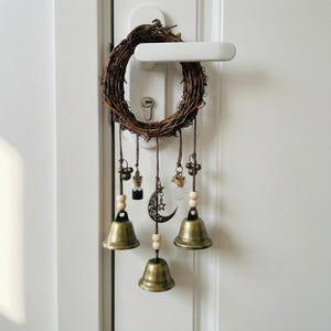 May include: A rustic wreath made of woven brown twigs, adorned with hanging bronze bells, small glass bottles, wooden beads, and a crescent moon charm. The wreath is suspended from a white door handle, creating a whimsical, decorative accent.