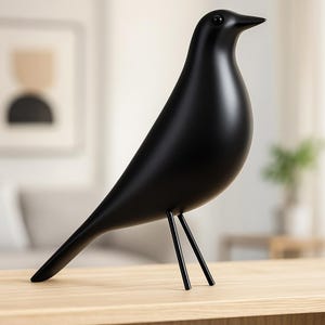 Könnte beinhalten: Eine matte, schwarze dekorative Vogelfigur mit einem schlanken, modernen Design. Der Vogel hat einen glatten, abgerundeten Körper und einen langen, spitzen Schnabel. Er steht auf drei dünnen, schwarzen Metallbeinen und steht auf einer hellen Holzoberfläche.