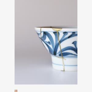 Può includere: Ciotola in ceramica bianca con motivi floreali blu e riparazioni kintsugi dorate. Il design della ciotola combina estetica tradizionale e moderna, mostrando la bellezza dell'imperfezione. Circa 15 cm di diametro.