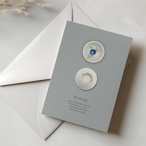 May include: A greeting card with two small, white ceramic dishes on a gray background. The top dish has a blue floral design. The card reads "Kintsugi" and includes a quote. A white envelope is partially visible.