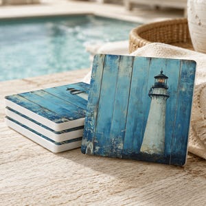 May include: A stack of square coasters featuring a printed image of a white lighthouse against a background of blue, weathered wooden planks. The design shows a tall lighthouse structure with a lantern room at the top, set against vertical boards.