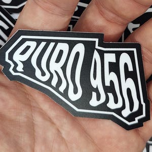 May include: A black and white sticker with the text "EURO 956" in a stylized font. The sticker has a unique shape and is held in a hand, with other similar stickers visible in the background.