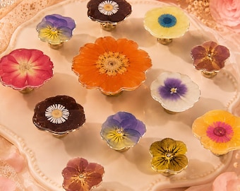 May include: A collection of decorative cabinet knobs with pressed flower designs. The knobs feature various flower types in shades of red, orange, yellow, purple, and brown, set on gold-toned bases. They are arranged on a white, scalloped-edge plate.