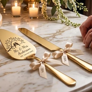 May include: A three-tiered white wedding cake with gold accents and decorative icing sits next to a gold cake server and knife set. The server is engraved with a tree and bird design and the names "Mr. & Mrs. Anderson".