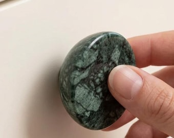 May include: A round cabinet knob made of dark green stone with natural white and black veining patterns. The knob has a smooth, polished surface and is attached to a plain, off-white drawer front, showcasing its circular shape and texture.