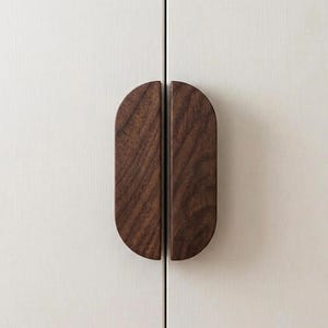 May include: Two semi-oval dark brown wooden cabinet handles mounted vertically on a light off-white cabinet door. The handles feature a visible wood grain pattern and are positioned side-by-side to form a complete oval shape when closed.