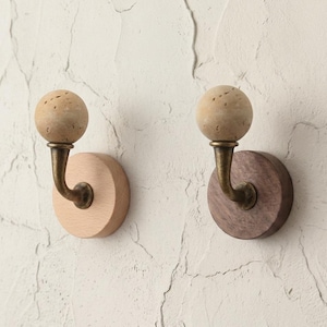 Puede incluir: Dos perchas de pared de madera. Una tiene una base de madera clara y la otra una base de madera oscura. Cada percha tiene un brazo curvo de color latón y una parte superior esférica, texturizada y de color beige.