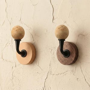 May include: Two wooden wall hooks. One hook has a light wood base and the other has a dark wood base. Each hook has a black metal curved arm and a spherical, textured, light brown top.