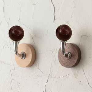 May include: Two wall-mounted coat hooks. Each hook features a dark red marble-like sphere atop a curved silver metal arm, mounted on a round wooden base. One base is light-colored wood, the other is dark wood.