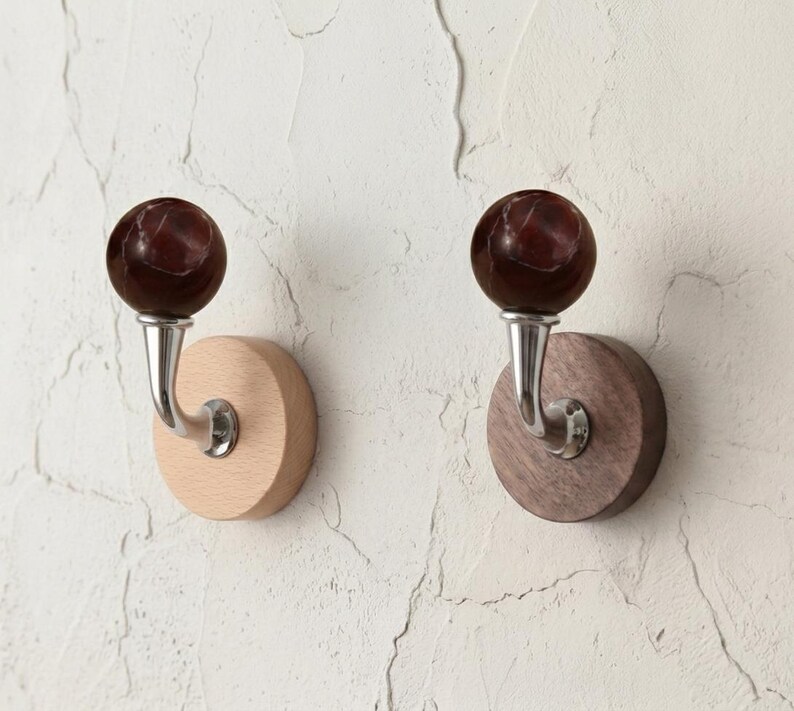 May include: Two wall-mounted coat hooks. Each hook features a dark red marble-like sphere atop a curved silver metal arm, mounted on a round wooden base. One base is light-colored wood, the other is dark wood.