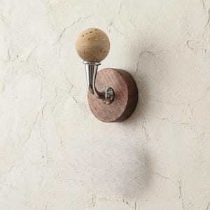 May include: A wall-mounted coat hook with a round, dark wood base and a curved metal arm. The hook features a spherical, light-colored, textured knob. The hook is mounted on a textured, off-white wall.