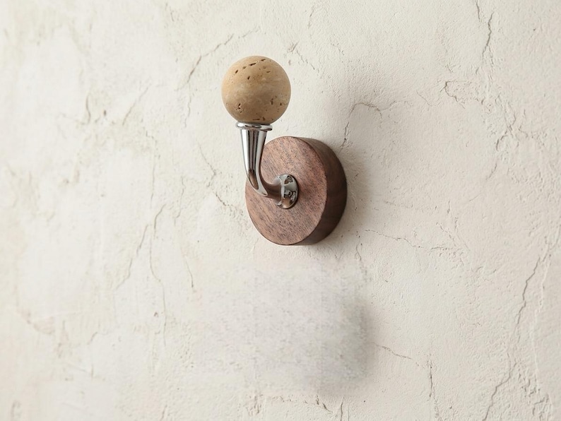 May include: A wall-mounted coat hook with a round, dark wood base and a curved metal arm. The hook features a spherical, light-colored, textured knob. The hook is mounted on a textured, off-white wall.