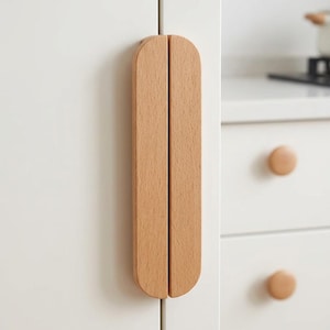 Puede incluir: Un tirador de armario de madera de color marrón claro con una forma vertical, alargada y semicircular. El tirador está montado en una puerta de armario blanca, con una textura de veta de madera natural lisa y una ranura vertical recta.