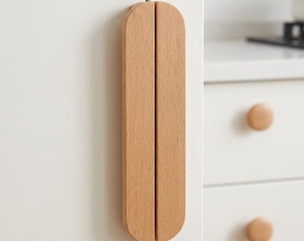 May include: A light brown wooden cabinet handle with a vertical, elongated, semi-circular shape. The handle is mounted on a white cabinet door, featuring a smooth, natural wood grain texture and a clean, straight vertical split down the center.