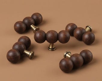 May include: Four cabinet pulls, each featuring three dark brown wooden spheres arranged in a horizontal row. Each pull is attached to a central brass-colored metal base that connects to the furniture surface, showcasing a natural wood grain texture.