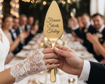 May include: A gold-colored cake server with the inscription "Forever Starts Today" is held by a couple at a wedding reception. The server has a decorative edge and a long handle. The bride wears a lace glove and a wedding ring.