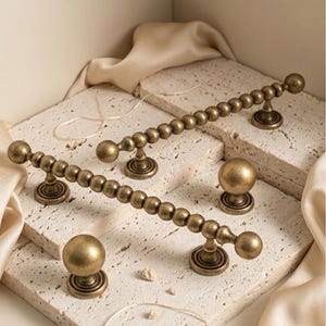 May include: Antique brass cabinet hardware set. The set includes two long beaded pulls, two round knobs, and two smaller pulls. The hardware has a vintage aesthetic and is displayed on a textured surface with a neutral background.