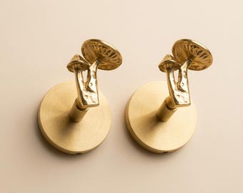 May include: Two matching wall hooks made of brushed gold-toned metal. Each hook features a circular base plate and a decorative arm shaped like a cluster of two mushrooms with textured, organic-looking stems and detailed, ribbed mushroom caps.