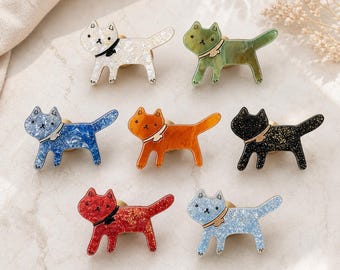 May include: A collection of seven cat-shaped pins in various colors including white, green, blue, orange, black, red, and light blue. Each pin features a glittery or marbled finish, a small collar, and a simple drawn face with ears and a tail.