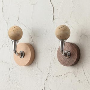 May include: Two wall hooks with a natural cork sphere top and a curved metal hook. One hook has a light wood base, the other a dark wood base. The hooks are mounted on a textured white wall.