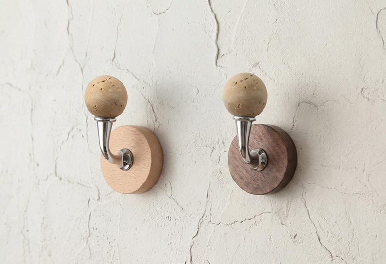 May include: Two wall hooks with a natural cork sphere top and a curved metal hook. One hook has a light wood base, the other a dark wood base. The hooks are mounted on a textured white wall.