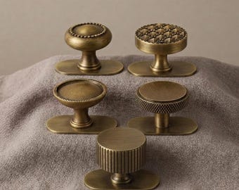 May include: Five antique brass cabinet knobs with varied textures and shapes, each mounted on an oval base plate. The collection includes designs with beaded edges, a pyramid grid pattern, a smooth circular top, a knurled edge, and vertical fluting.