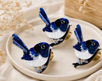 May include: Three decorative cabinet knobs shaped like birds, featuring glossy blue, black, and white acrylic layers with a metallic gold-toned base. Each bird figure is depicted in a perched position with a long tail, dark eye, and detailed wing patterns.