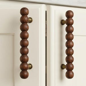 May include: Two vertical cabinet handles made of dark brown wood, each featuring a series of eight spherical beads stacked in a column. The wooden beads are attached to the cream-colored cabinet doors by two small, cylindrical brass-toned metal mounts.