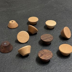 May include: A collection of eleven small, conical wooden knobs in varying shades of light and dark brown. Each knob features a smooth, rounded top surface and a tapered base, with some showing visible wood grain patterns and metal hardware inserts.