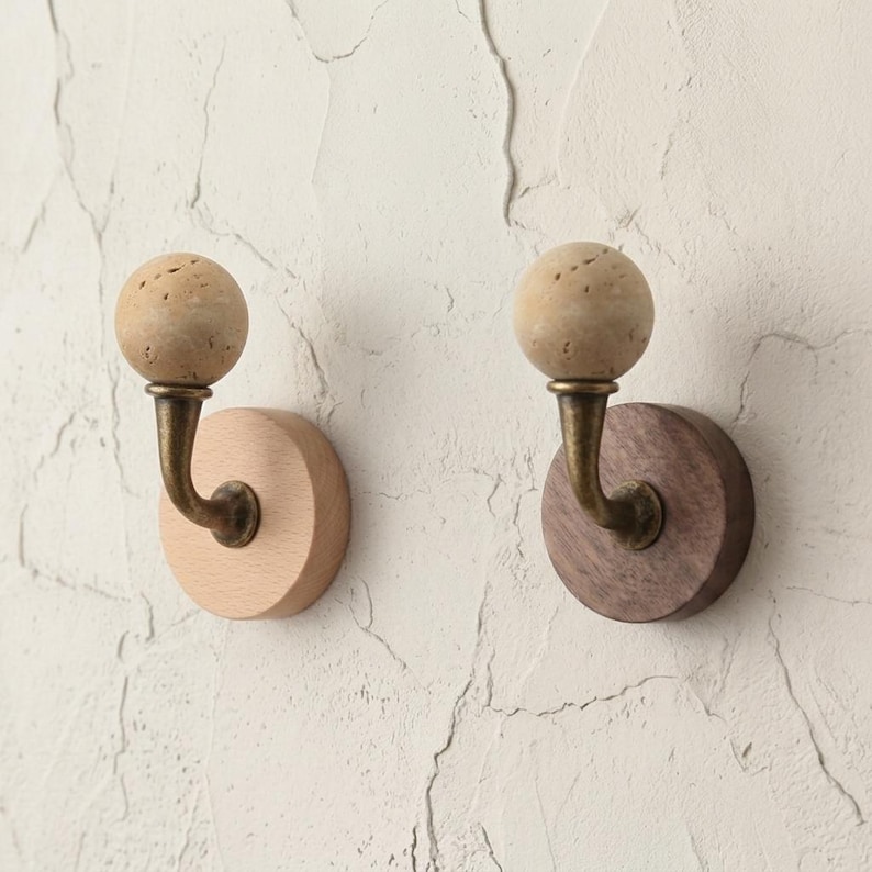 May include: Two wooden wall hooks. One hook has a light wood base and the other has a dark wood base. Each hook has a brass-colored curved arm and a spherical, textured, beige-colored top.