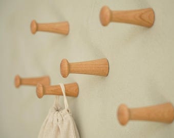 May include: A set of six light brown wooden wall hooks with a smooth, tapered cylindrical shape and a rounded knob end. One of the hooks holds a small, beige fabric drawstring pouch with a simple cord closure, mounted on a pale green wall.