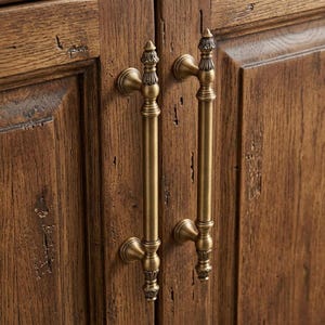 May include: Two vertical brass cabinet handles with a cylindrical shaft and decorative finials at the top and bottom, mounted on a pair of dark brown wooden cabinet doors featuring a visible wood grain and a distressed, textured surface finish.