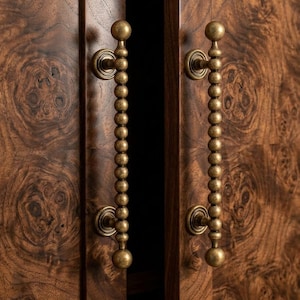 Puede incluir: Primer plano de un armario de madera con dos tiradores de cuentas de latón. Las puertas del armario son de color marrón oscuro con un patrón de vetas de madera único. Los tiradores son ornamentados, con una bola grande en la parte superior e inferior.
